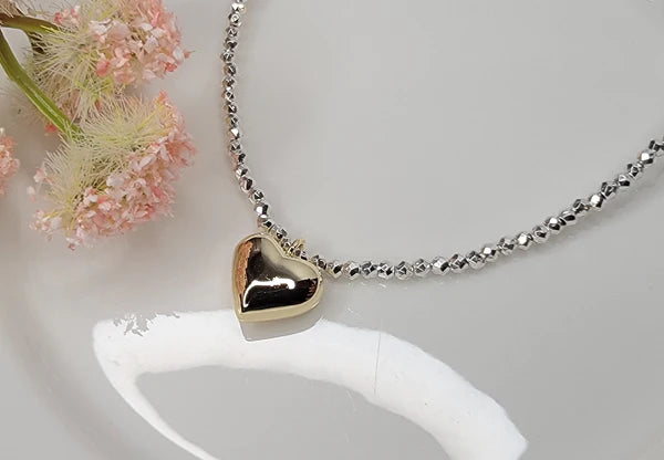 Heart-shaped pendant on a silver beaded chain against a white background with pink flowers.
