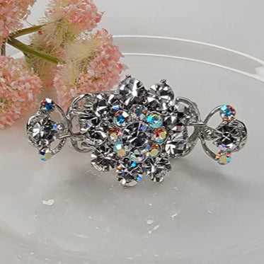Silver hair clip with crystal details on a white surface with pink flowers in the background